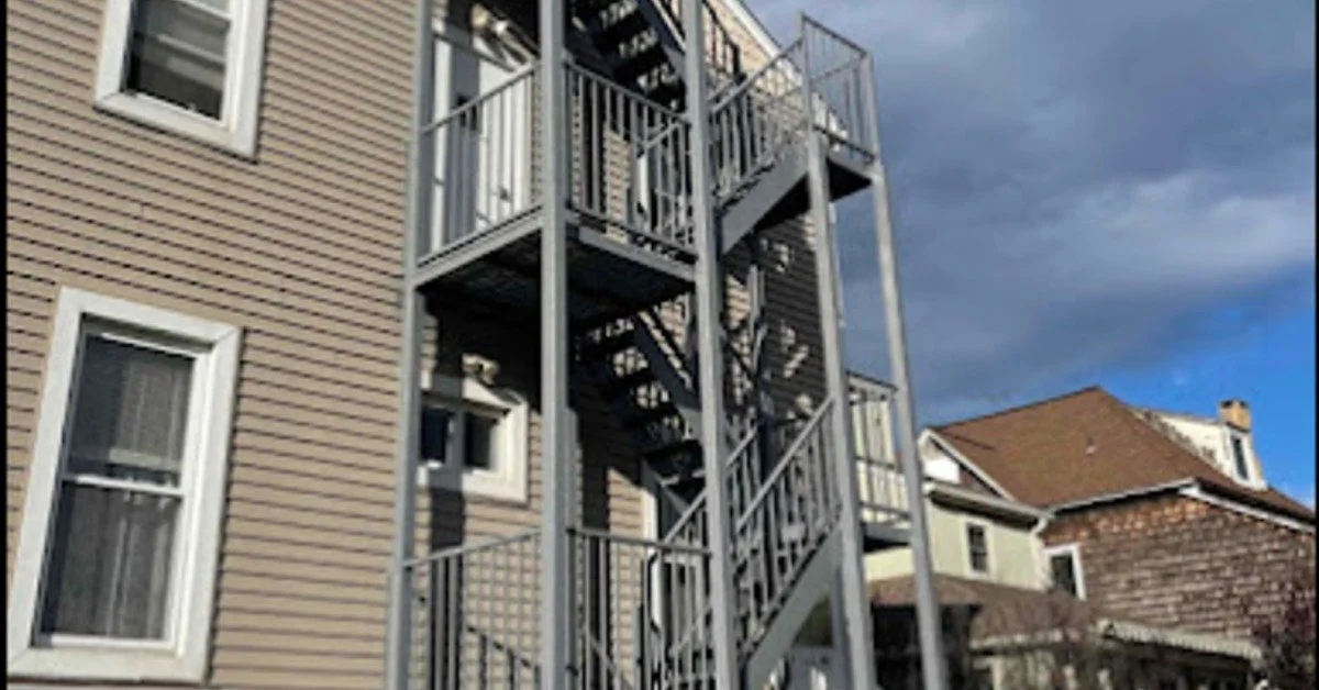 A beige apartment building with white-framed windows and a gray metal fire escape under cloudy blue sky.
