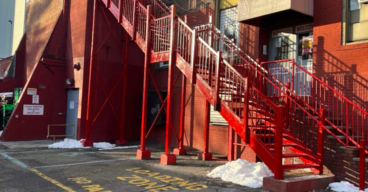 Red exterior fire escape beside a brick building with multiple stairways, parking lot, and entrance below.