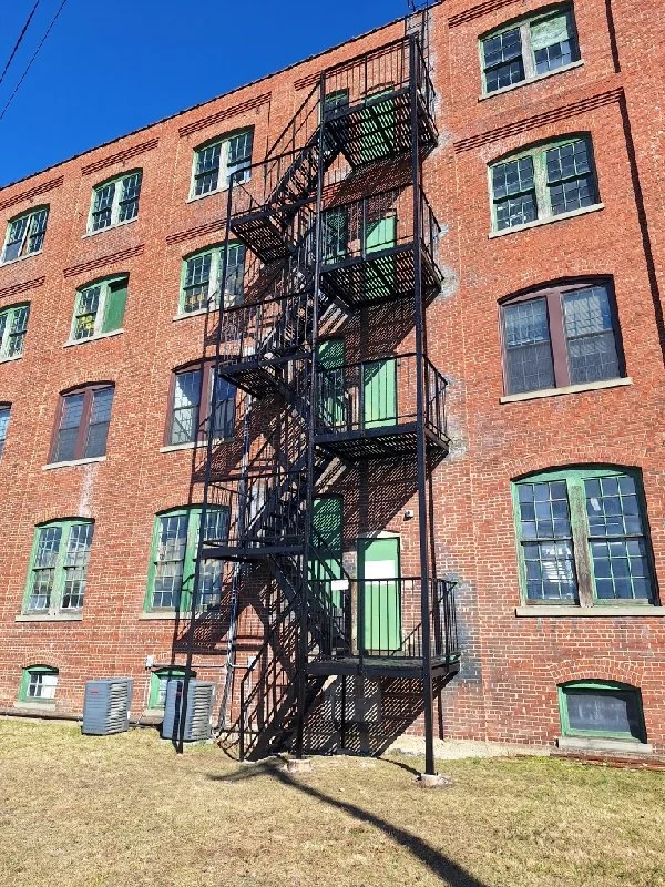 Exterior fire escape with stacked platforms and stairs rises along a brick building under a clear blue sky.
