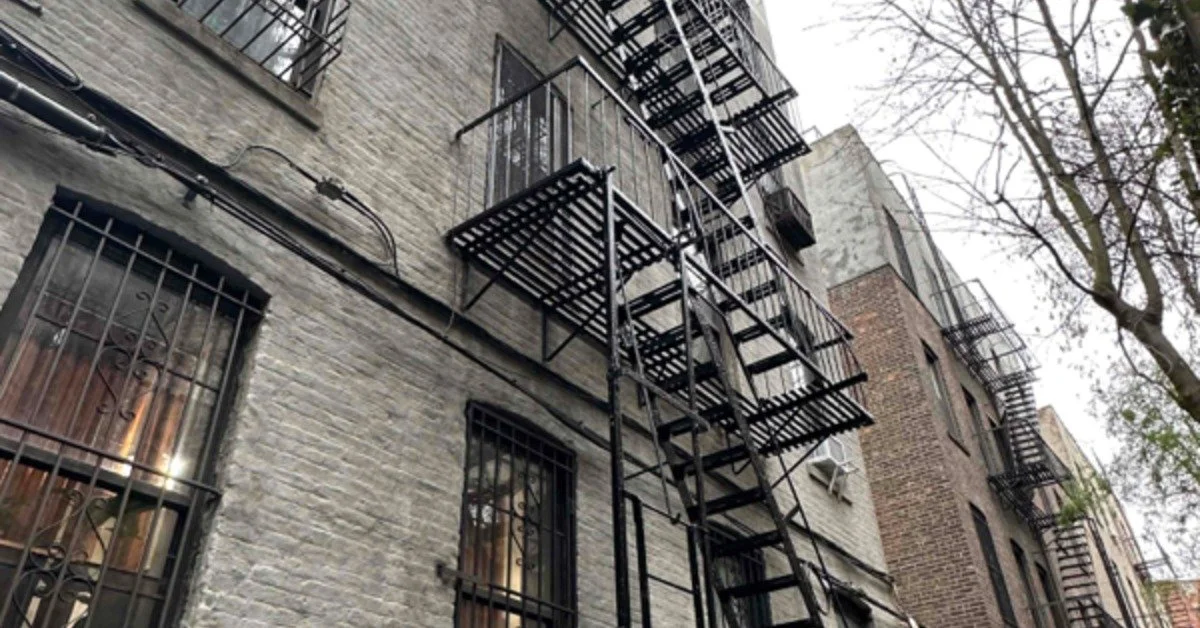 A gray brick apartment building with barred windows and black metal fire escapes beside bare tree branches.