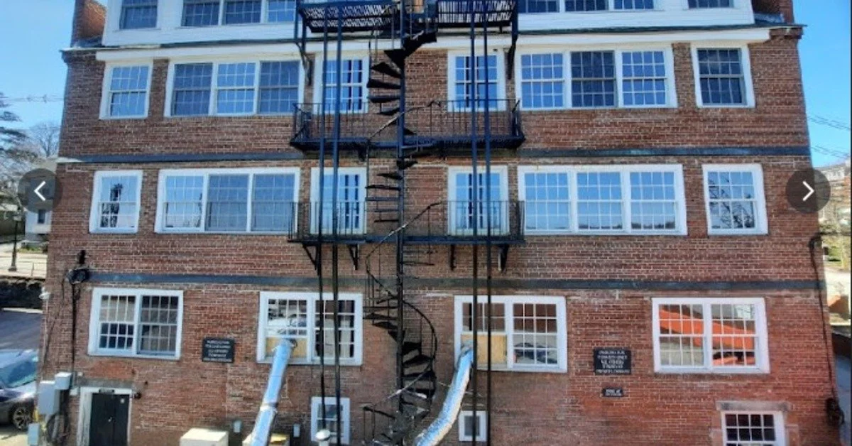 Black spiral fire escape climbs the center of a brick building facade with many windows and basement utilities.