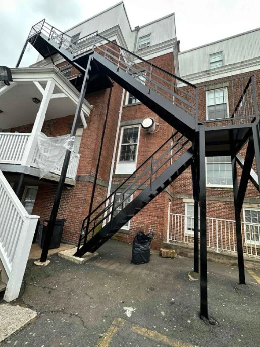 Black metal fire escape with multiple stair flights and platforms rises along the exterior of a red brick building.