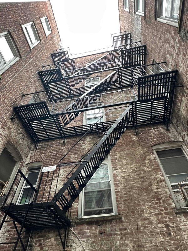 Upward view between brick buildings shows stacked black metal fire escape platforms, railings, and stairs.