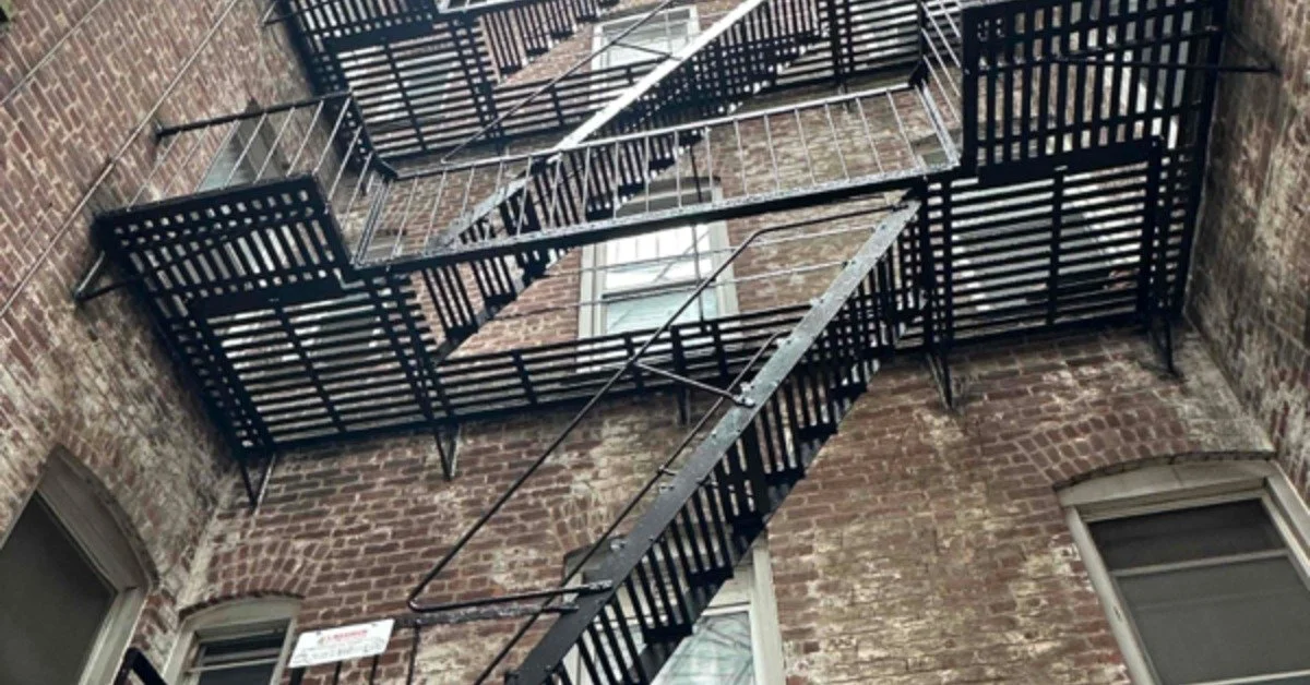Black metal fire escape zigzags between brick apartment walls, viewed upward through a narrow courtyard space.