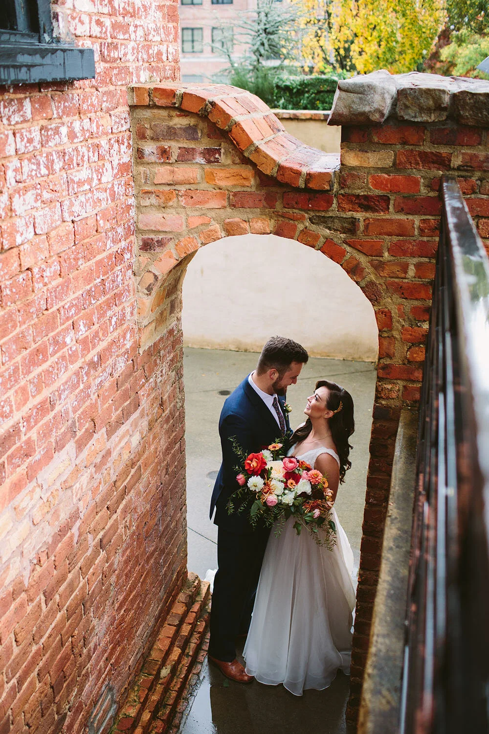 Penny_Bride_Groom_Smiling_Holding_Bridal_Bouquet_Under_Brick_Arch.jpg