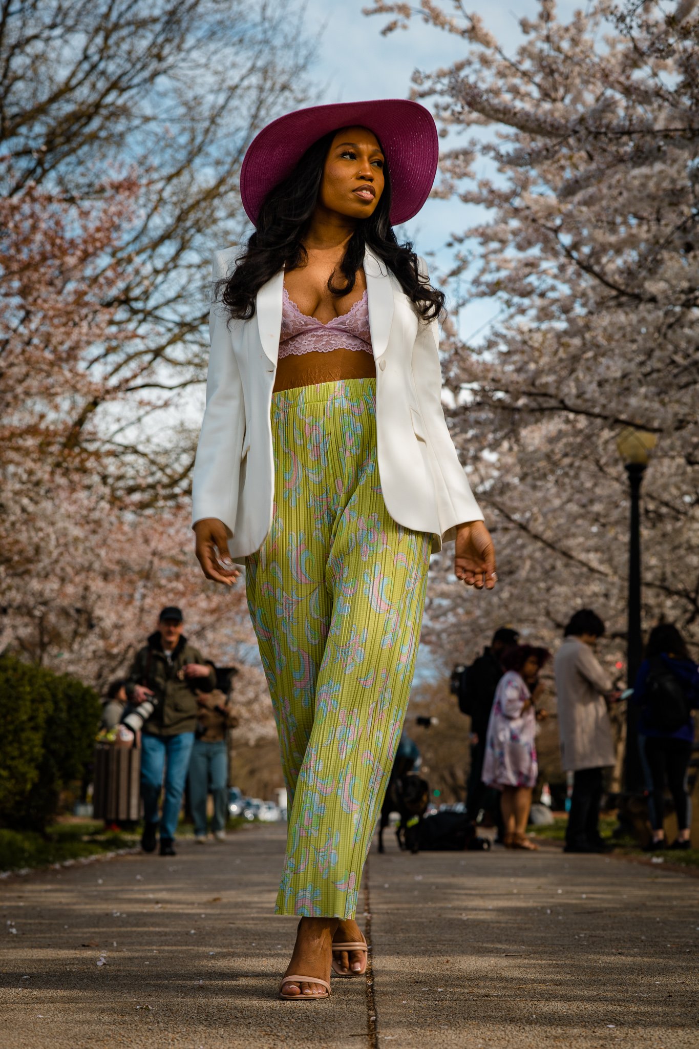 A woman walking outdoors in a park with cherry blossom trees in bloom, wearing a purple wide-brimmed hat, a white blazer, a lace pink top, yellow patterned pants, and clear high-heeled shoes.