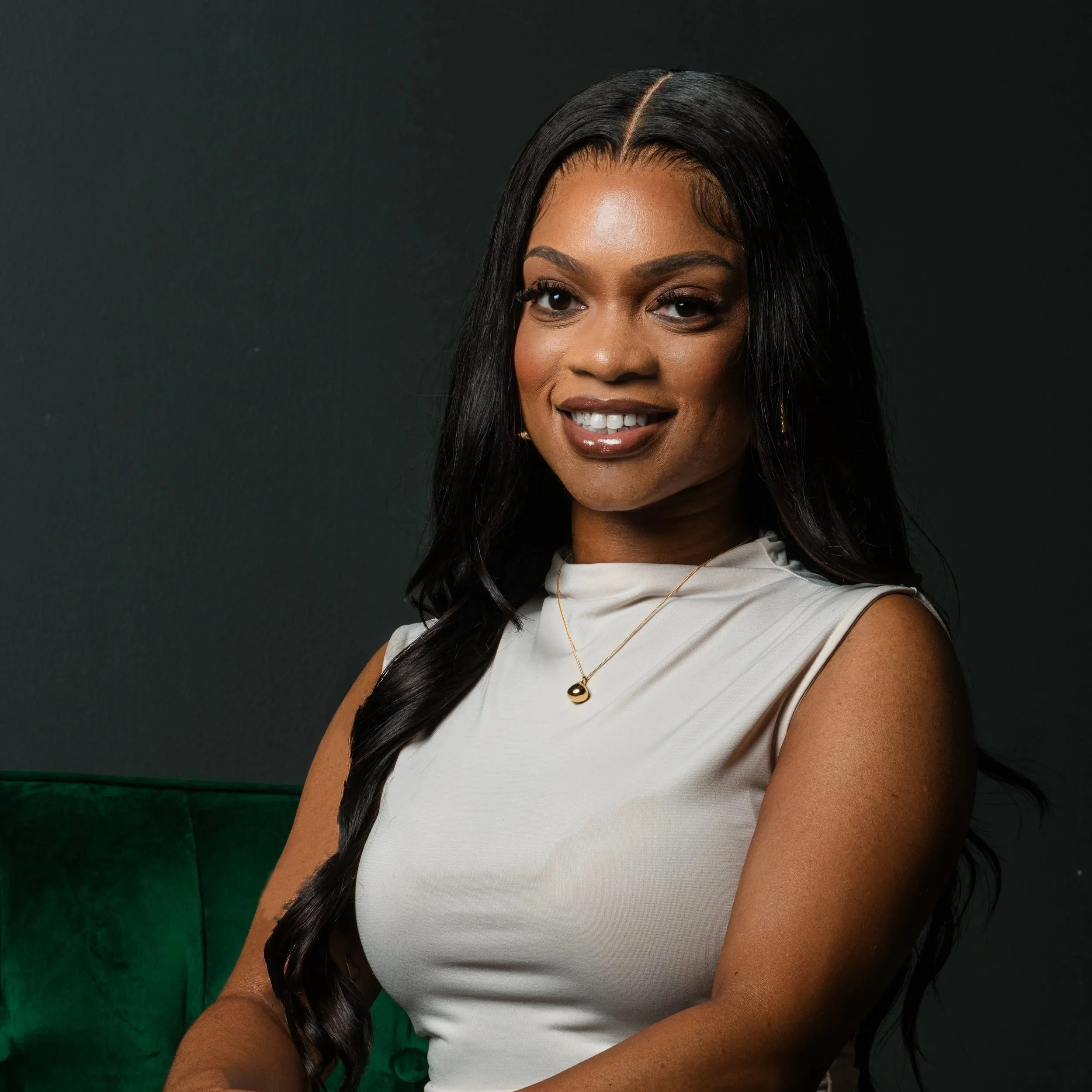 Portrait of a smiling woman with long black hair, wearing a white sleeveless top and gold jewelry, sitting on a green seat against a dark background.