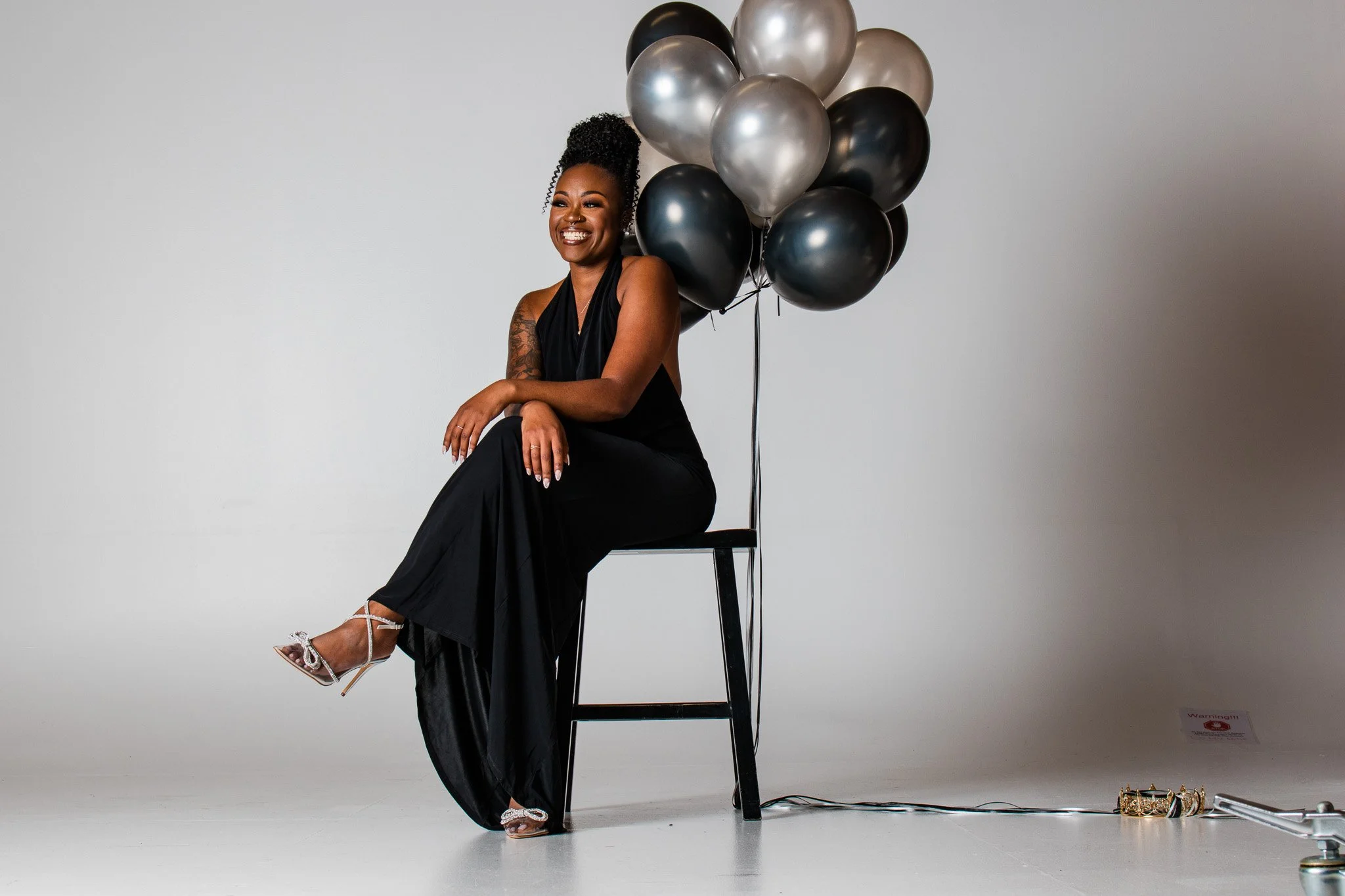 A woman sitting on a black chair in a white studio, smiling, wearing a black dress and heels, with a bunch of black, silver, and gray balloons behind her.
