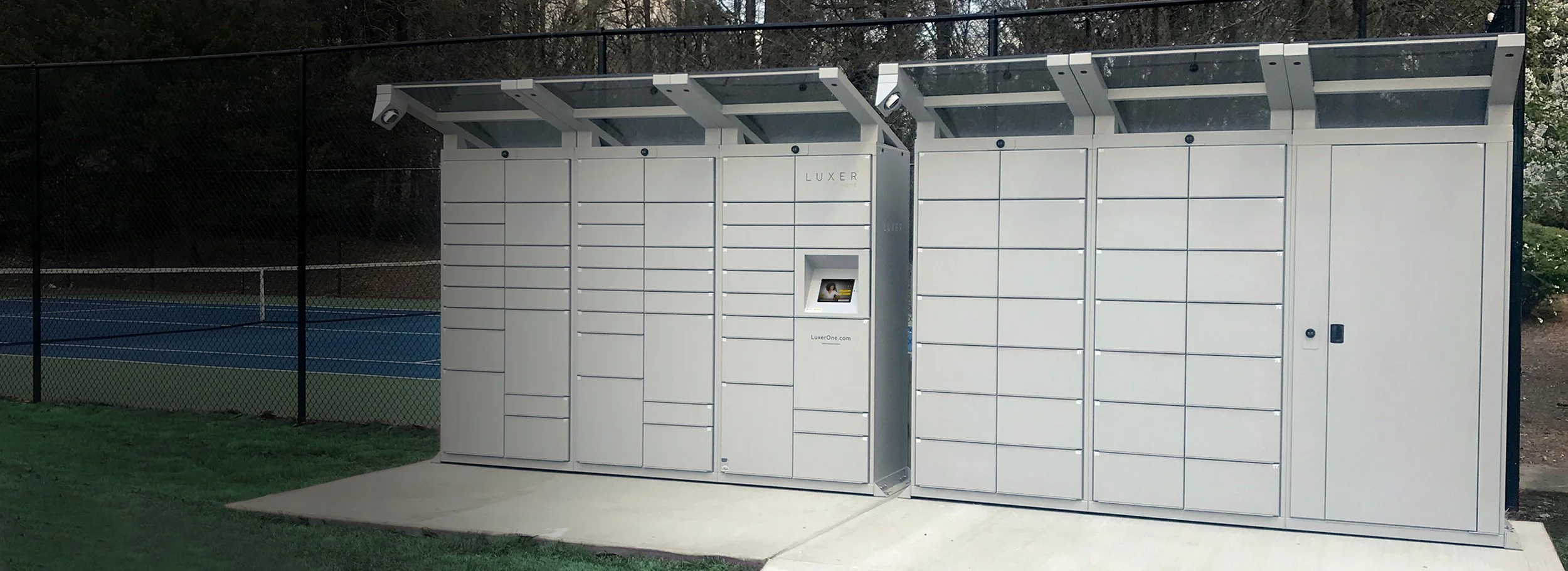 Two silver outdoor Luxer One locker banks with canopies next to a tennis court