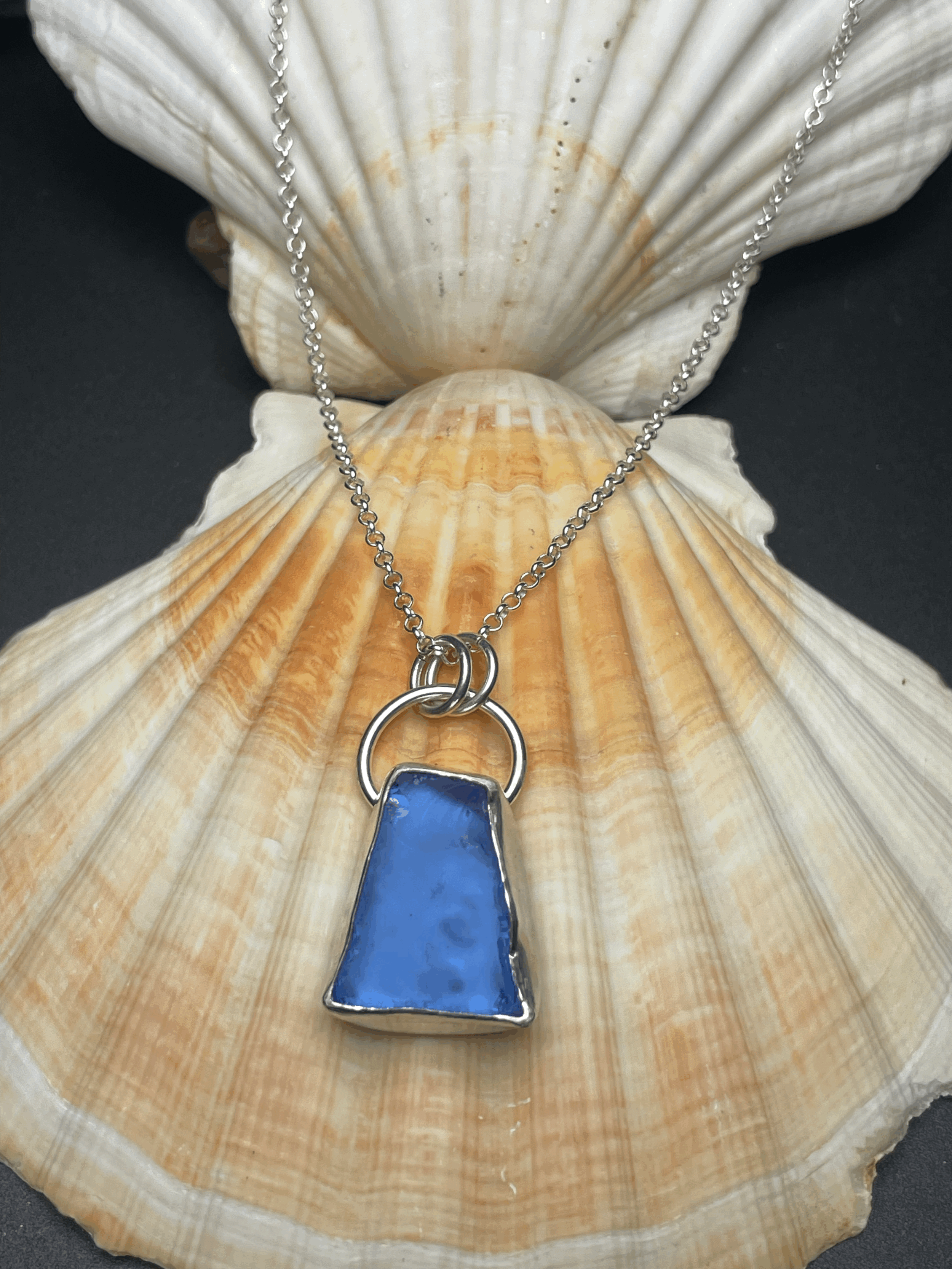 Silver necklace with a blue trapezoid-shaped pendant resting on a large seashell, black background.