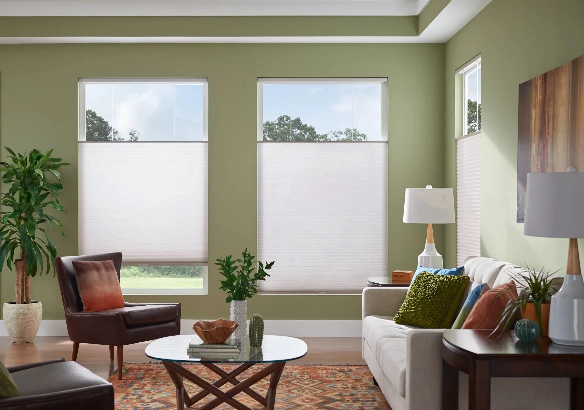 Living room with green walls, large windows with blinds, a white sofa with colorful pillows, a brown leather chair with an orange pillow, side tables with lamps, potted plants, and a patterned rug.