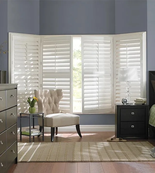 A cozy bedroom corner with a large window covered by white shutters, a beige tufted armchair, a small side table with a green plant, a black dresser on the left, and a black nightstand with a white lamp and books on the right. Light streams through the shutters onto the wooden floor and striped rug.