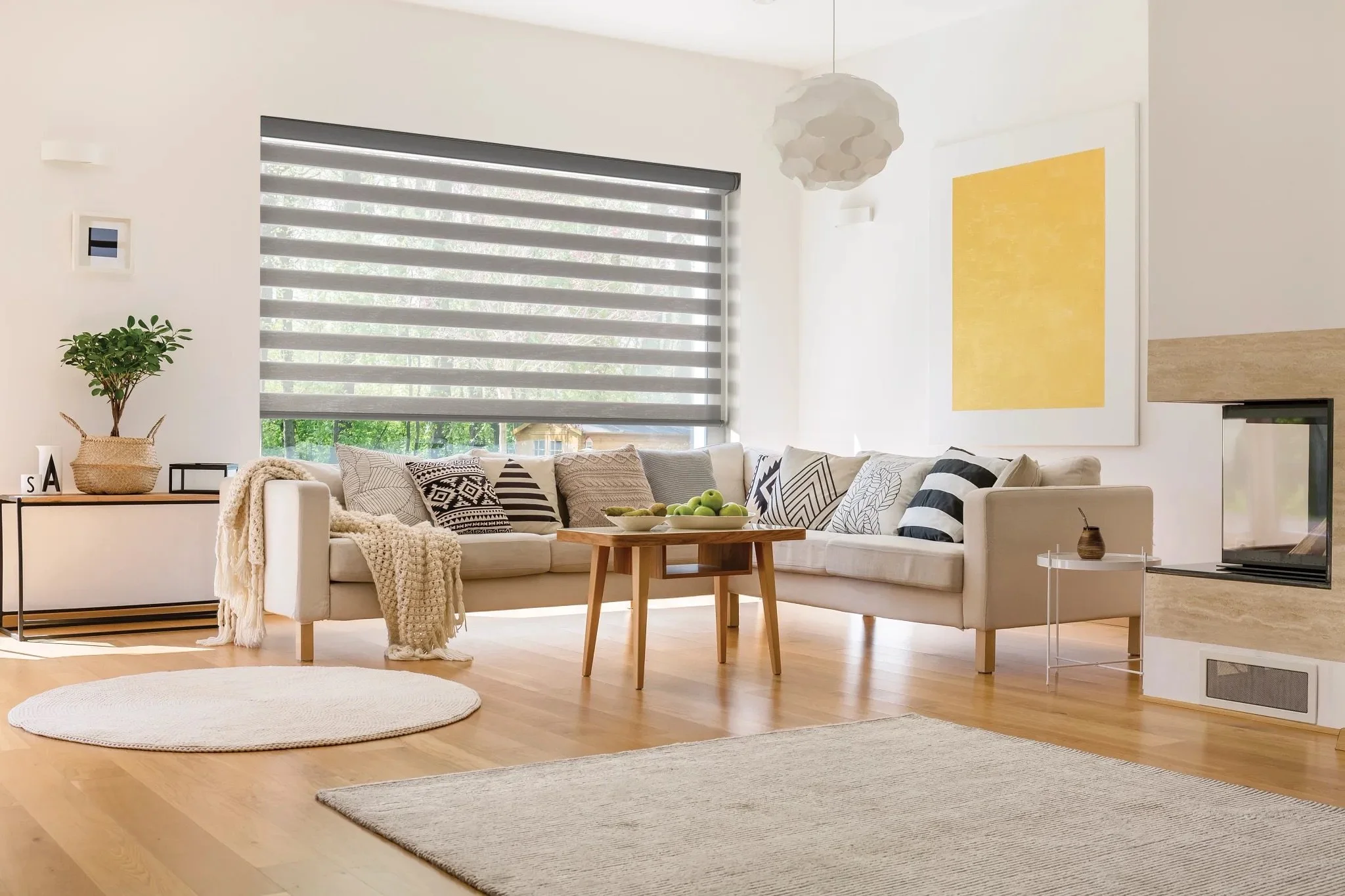 Bright and airy living room with white walls, large window with striped blinds, beige sectional sofa with black and white patterned pillows, round wooden coffee table with green apples, light wood flooring, abstract yellow painting on wall, potted plant, and modern decor.