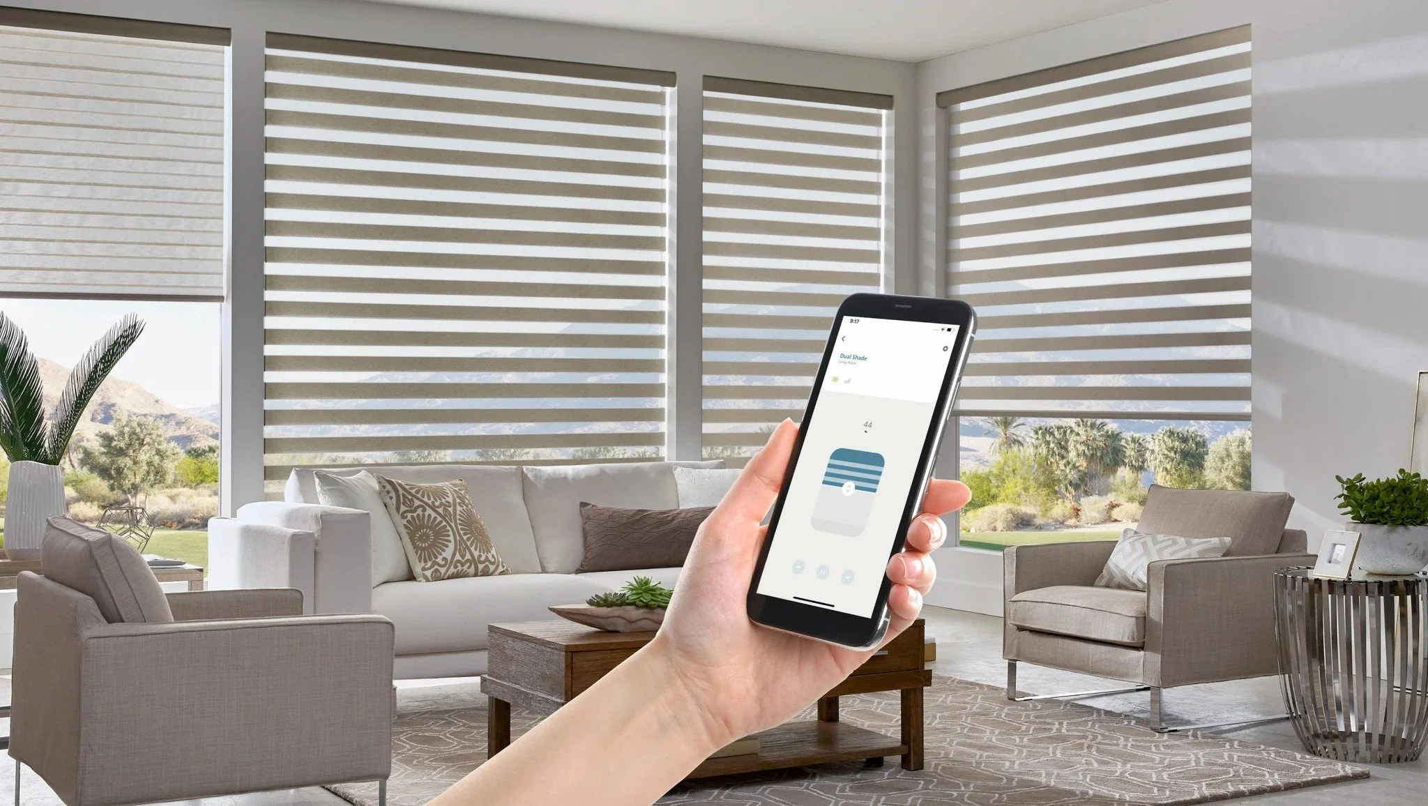 A living room with large windows featuring zebra blinds, a white sofa with patterned and plain pillows, armchairs, a coffee table, potted plants, and a person holding a smartphone controlling smart blinds.