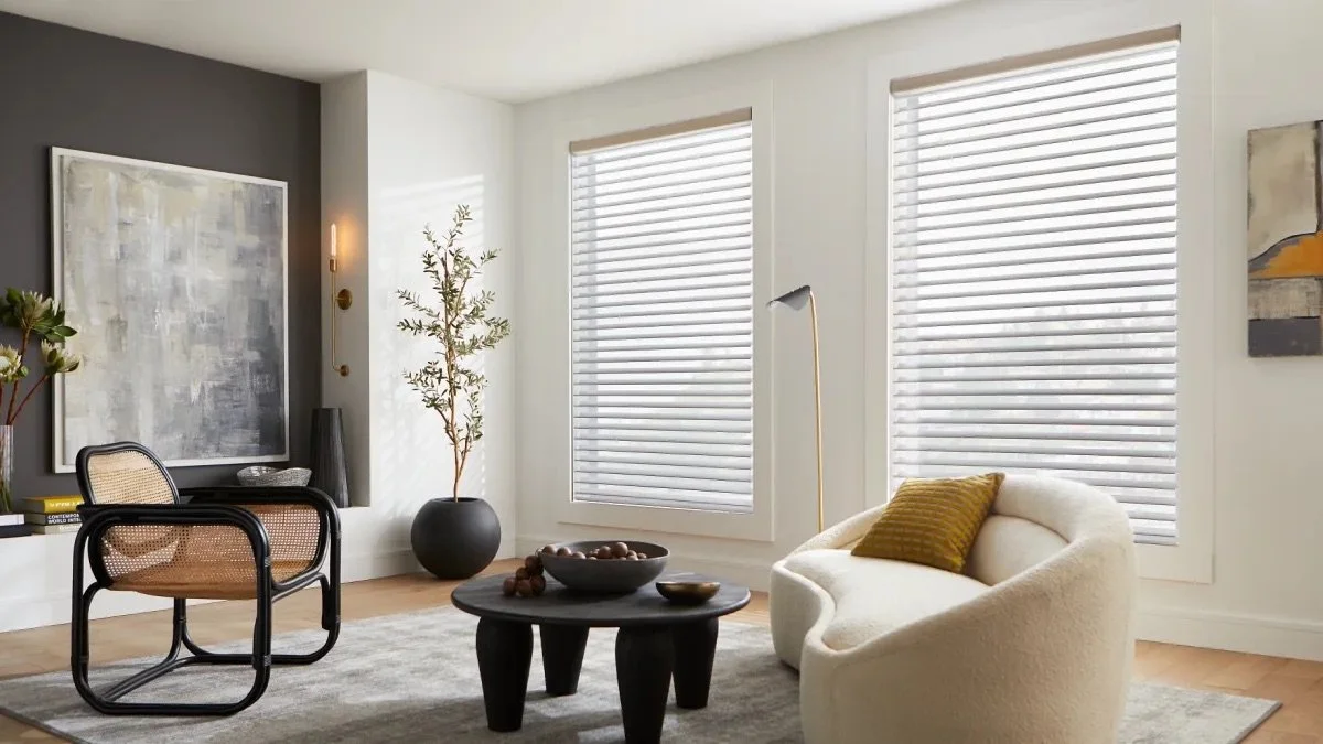 Modern living room with white walls, two large windows with blinds, a cream curved armchair with a yellow pillow, a black coffee table with bowls, a black-and-beige chair, an abstract painting, potted plants, a tall floor lamp, and a gray area rug.