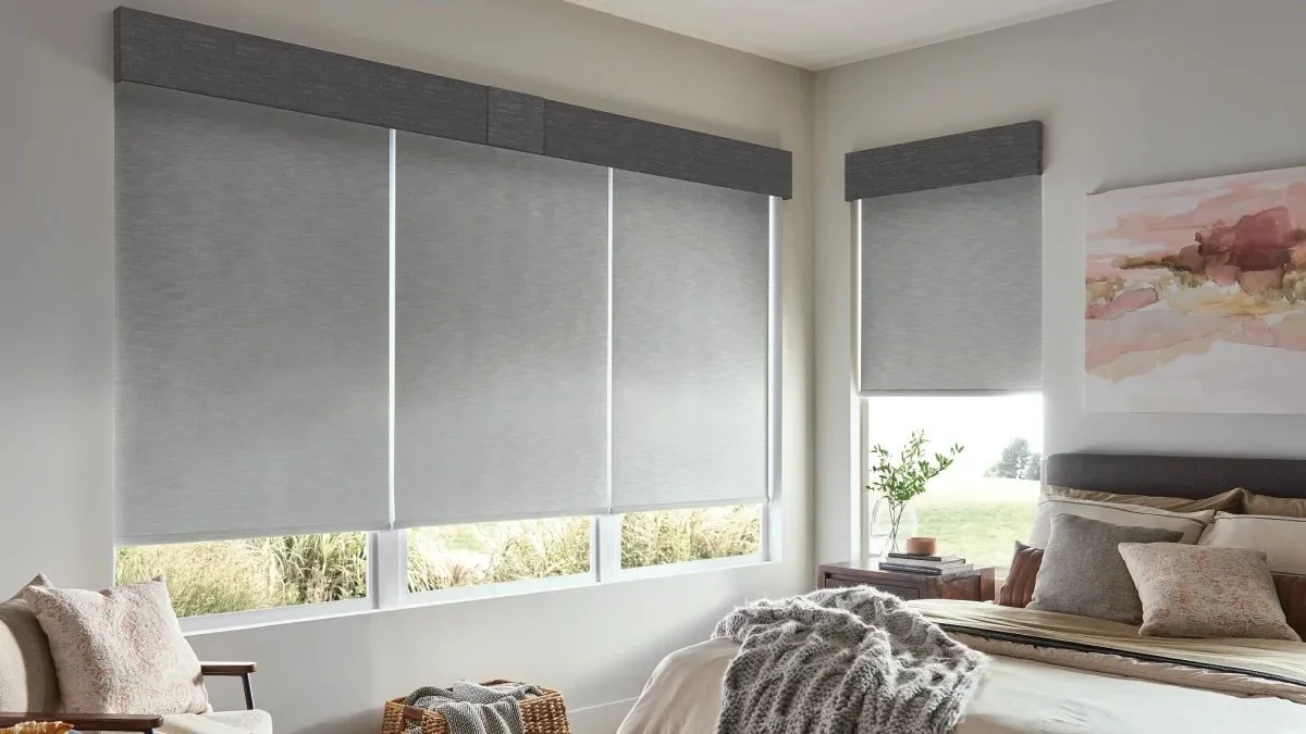 A bedroom with large windows covered by grey roller blinds, a bed with neutral-colored pillows and a blanket, a side table with a plant, and abstract wall art.
