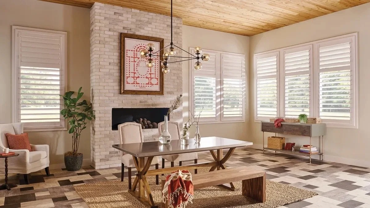 Bright living room with a white brick fireplace, large windows with white shutters, a wooden ceiling, a dining table with chairs, and decorative vases.