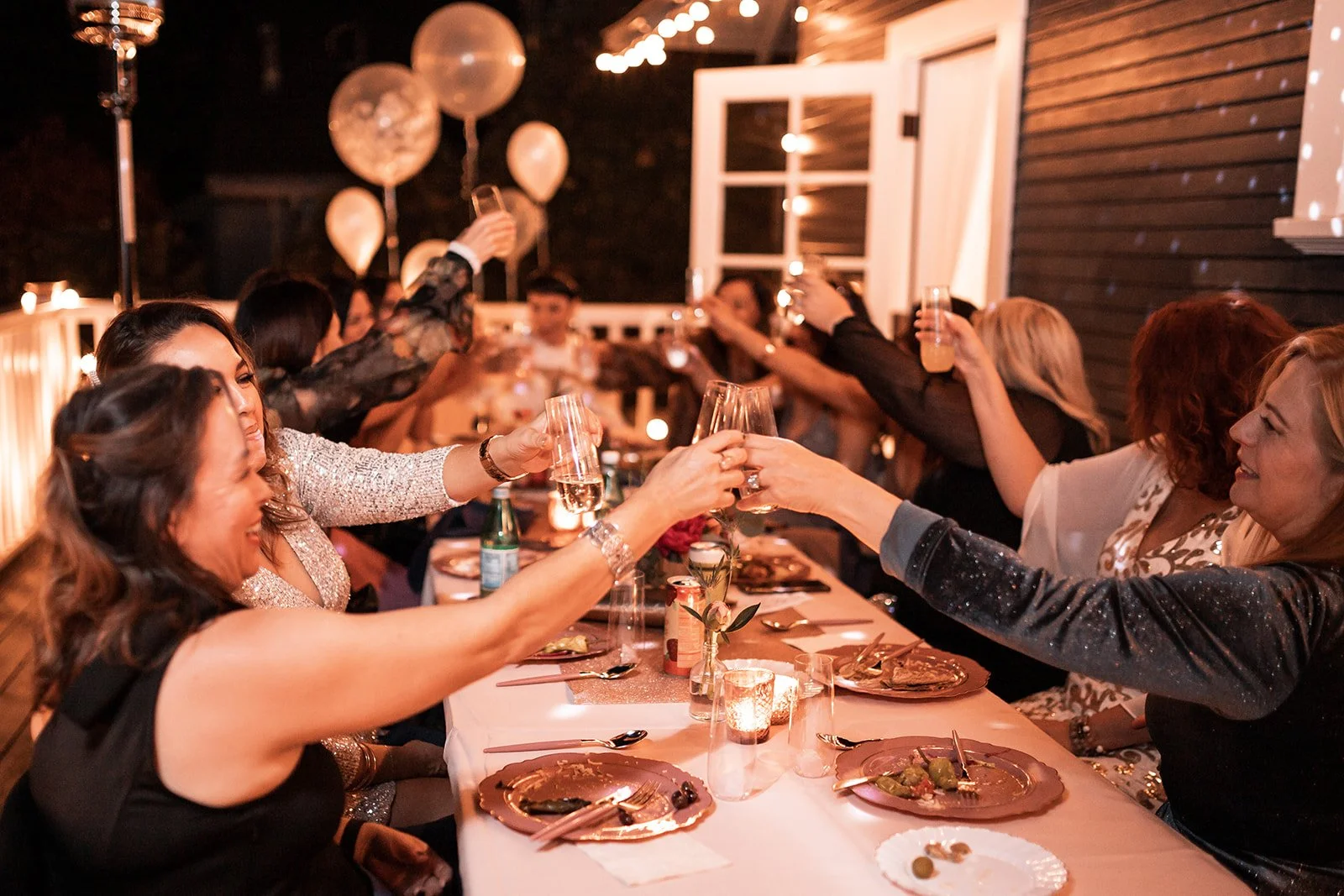 People at a dinner party raising glasses for a toast, seated around a table with plates, drinks, and candles, in a decorated outdoor setting at night.