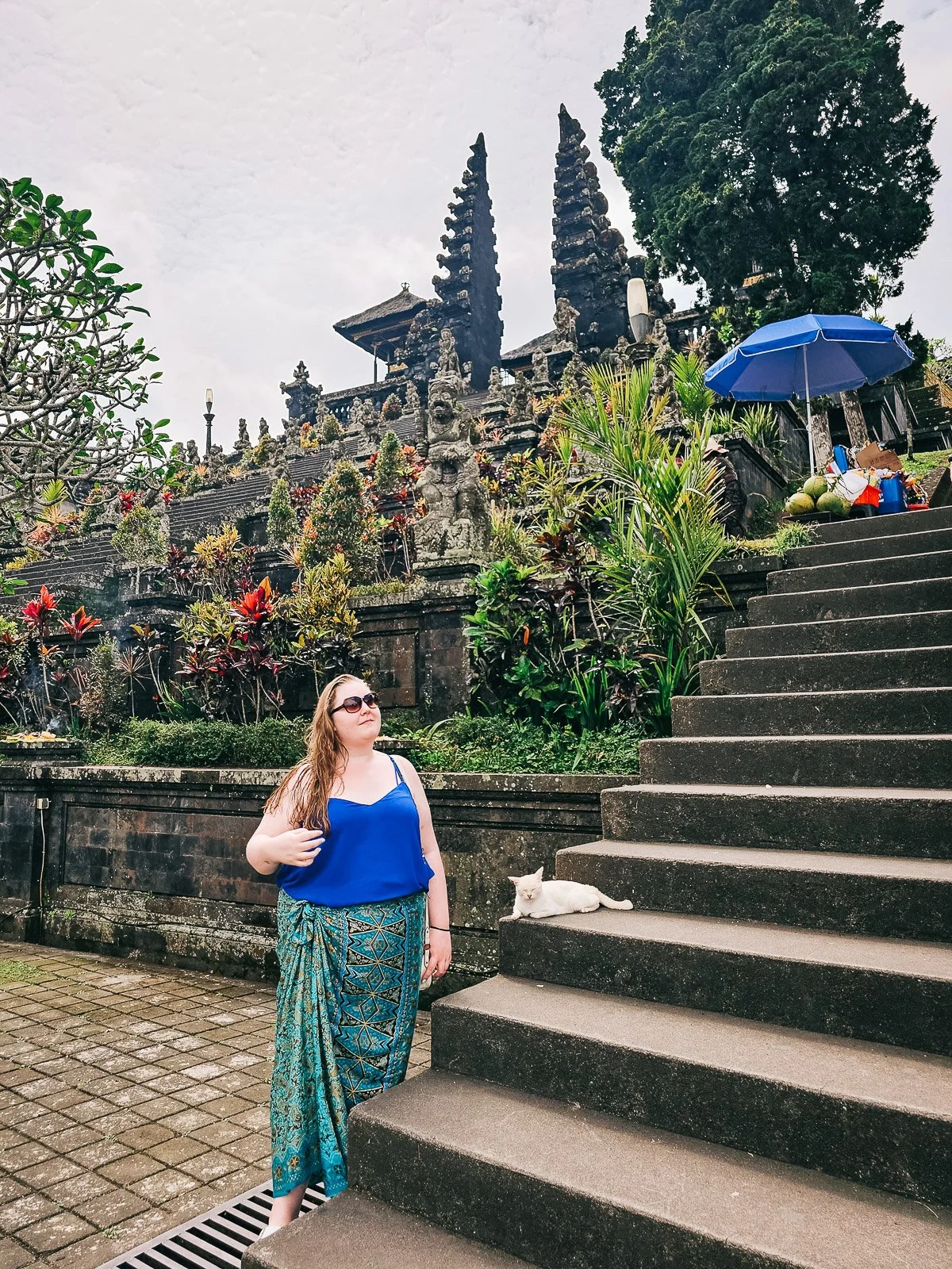 Traveler wearing a sarong at a Balinese temple in Indonesia with stone temple gates and tropical plants behind her.