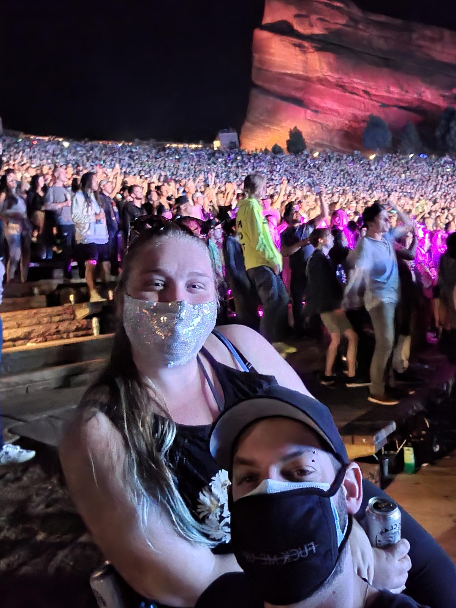 a woman and a man posing at red rocks amphitheater during a concert with a huge crowd behind them and a large rock formation