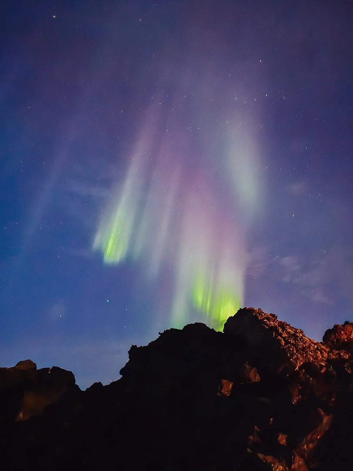 Green and purple Northern Lights above dark volcanic rocks in Iceland