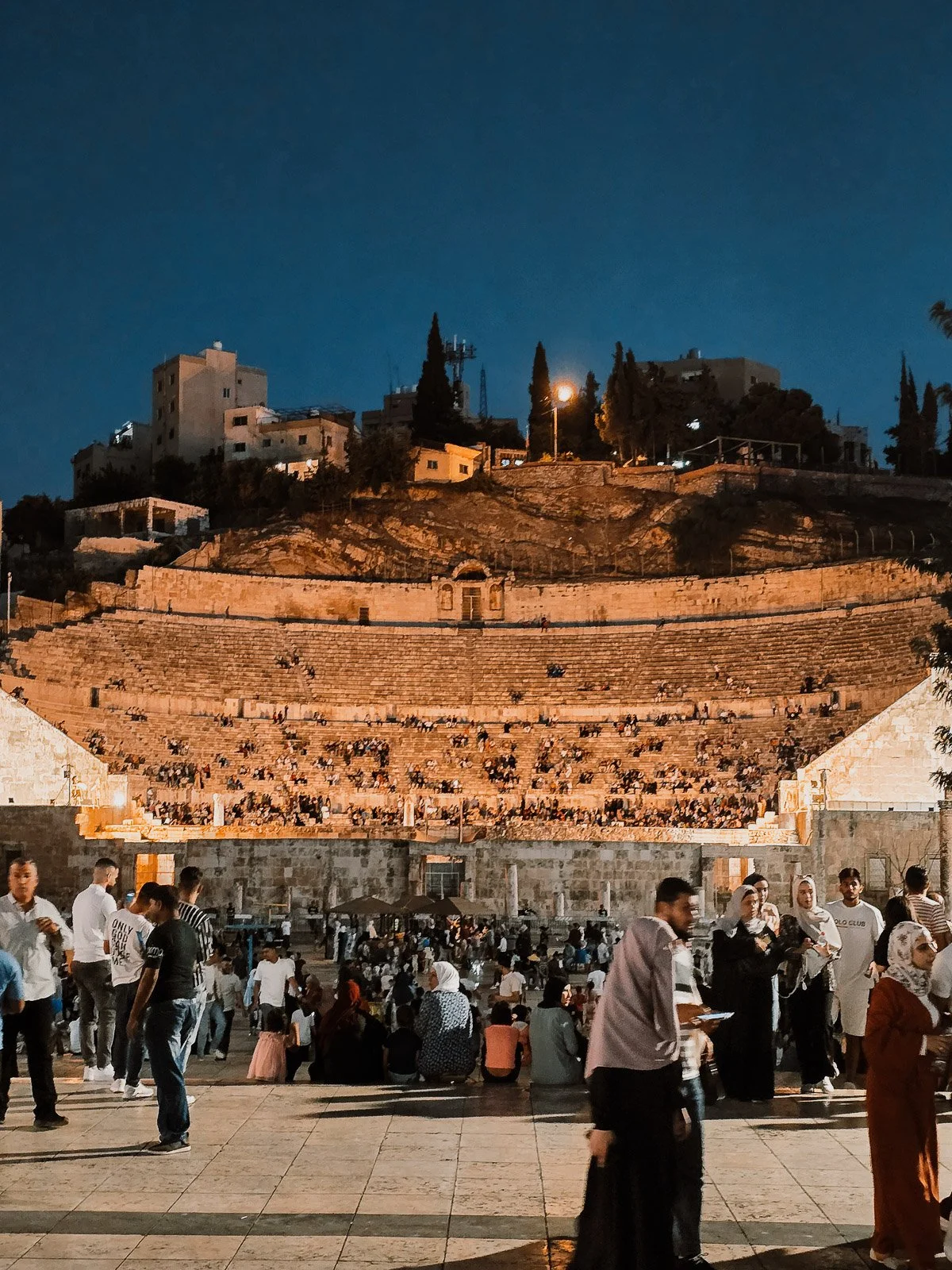 Crowds gathered at the Roman Theater in Amman, Jordan, illuminated at night with hillside buildings in the background.