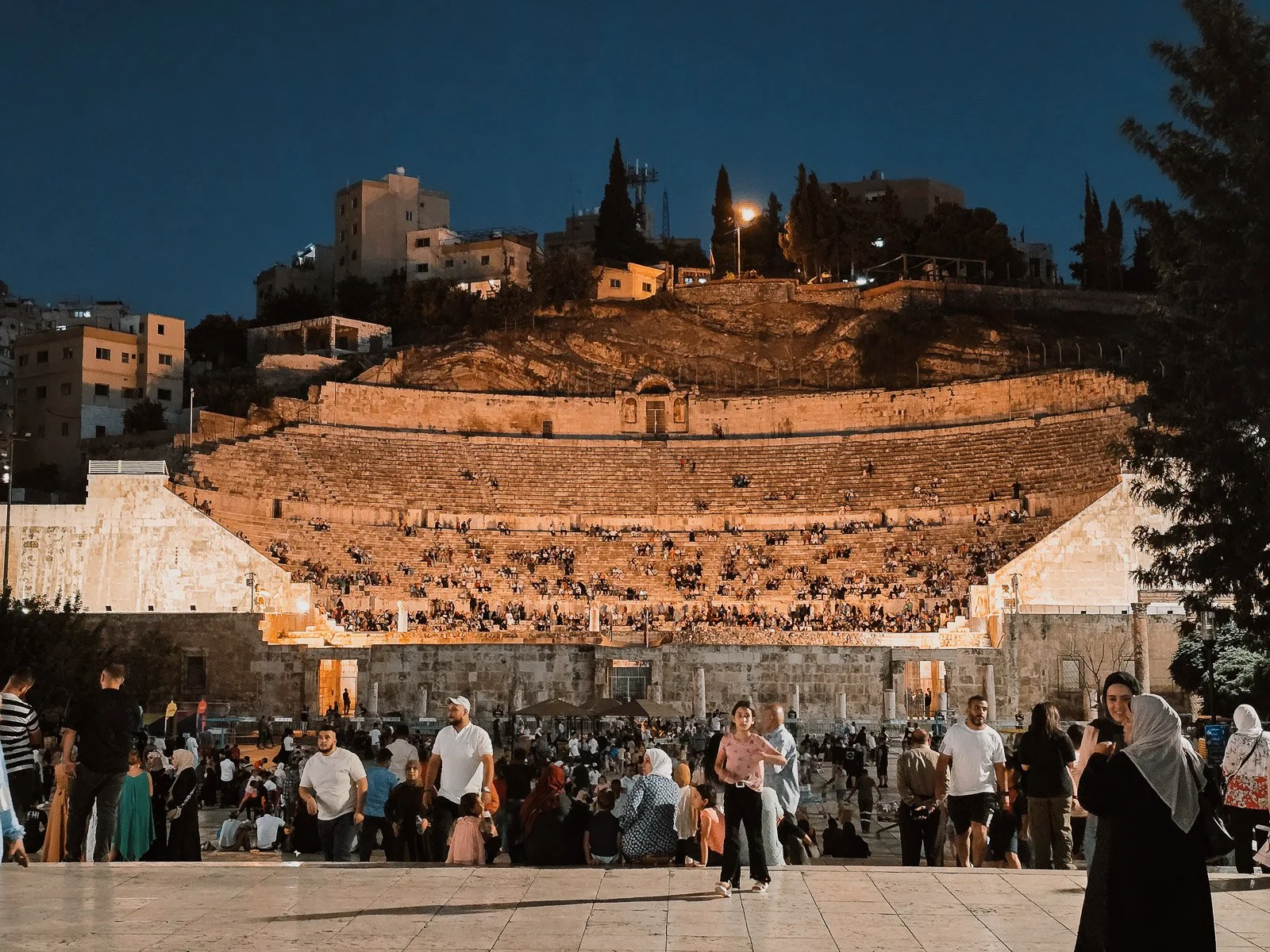 Crowds gathered in front of the Roman Theater in Amman, Jordan, illuminated at night beneath hillside buildings.
