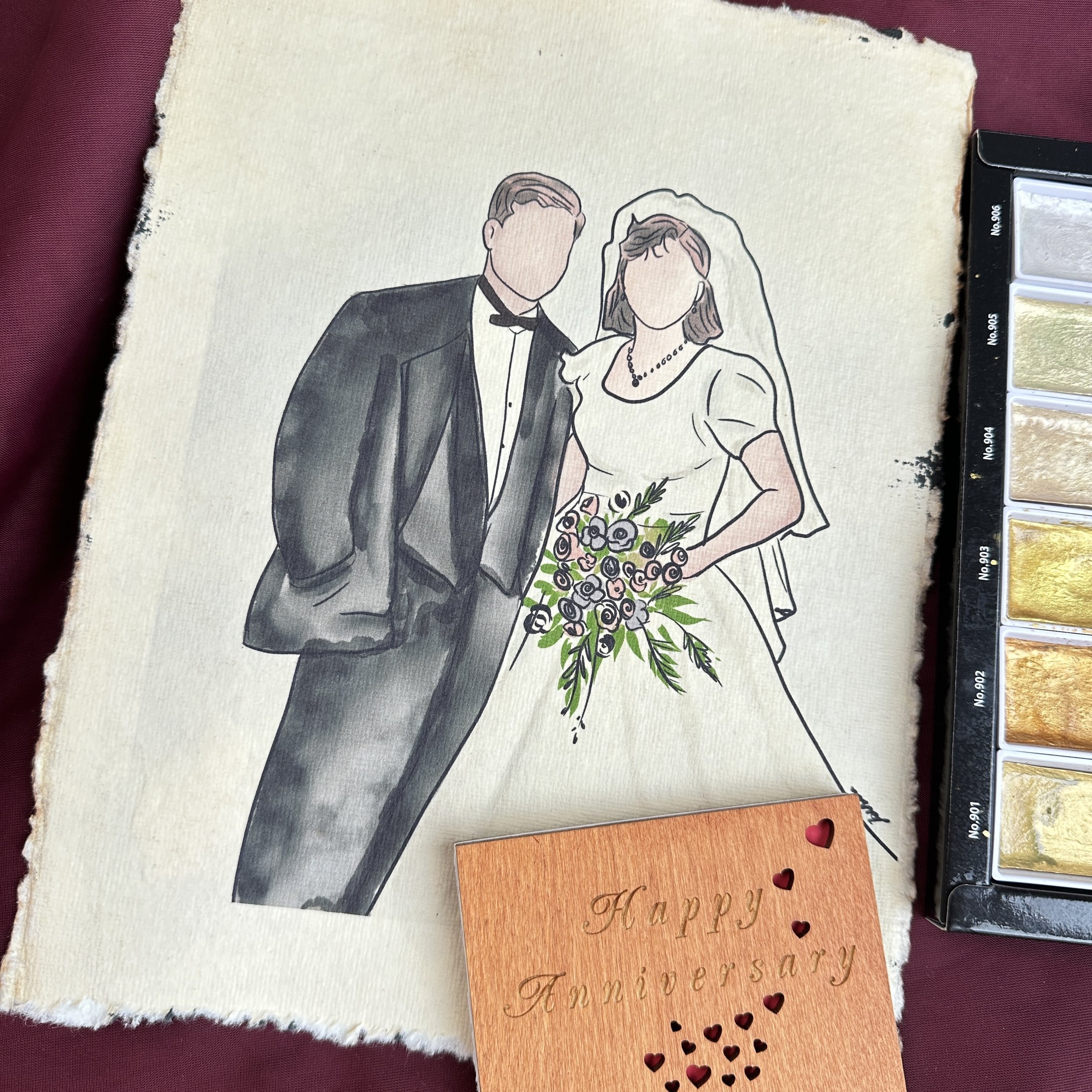 Handmade anniversary card featuring a watercolor illustration of a couple in wedding attire, with a bride holding a bouquet of flowers, on a textured piece of paper, next to a small orange card with the words "Happy Anniversary" and heart cutouts.