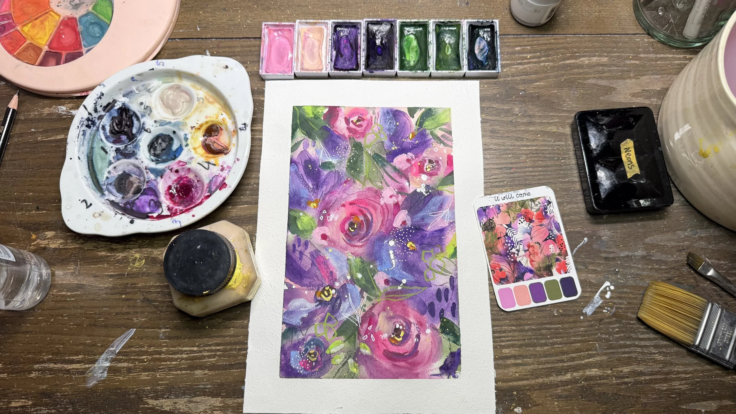 A watercolor painting of vibrant pink, purple, and green flowers on watercolor paper, surrounded by watercolor paints, brushes, and art supplies on a wooden table.