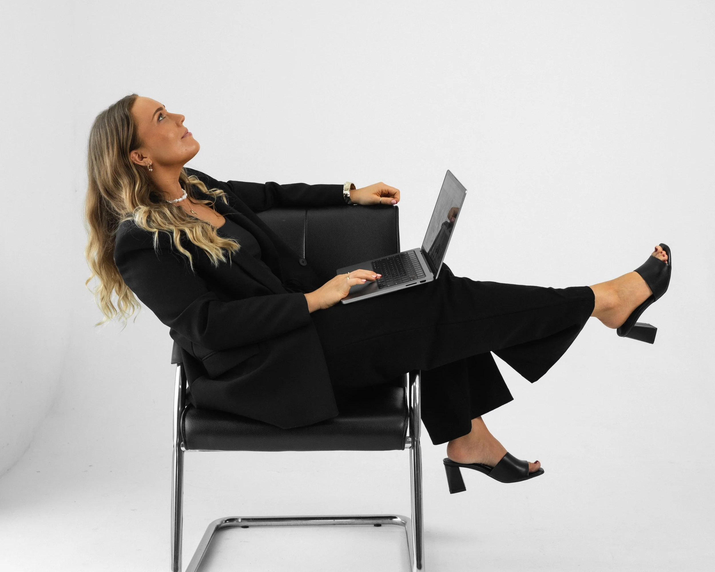 A woman dressed in a black suit sitting on a chair with her legs extended and crossed, working on a laptop, against a plain white background.
