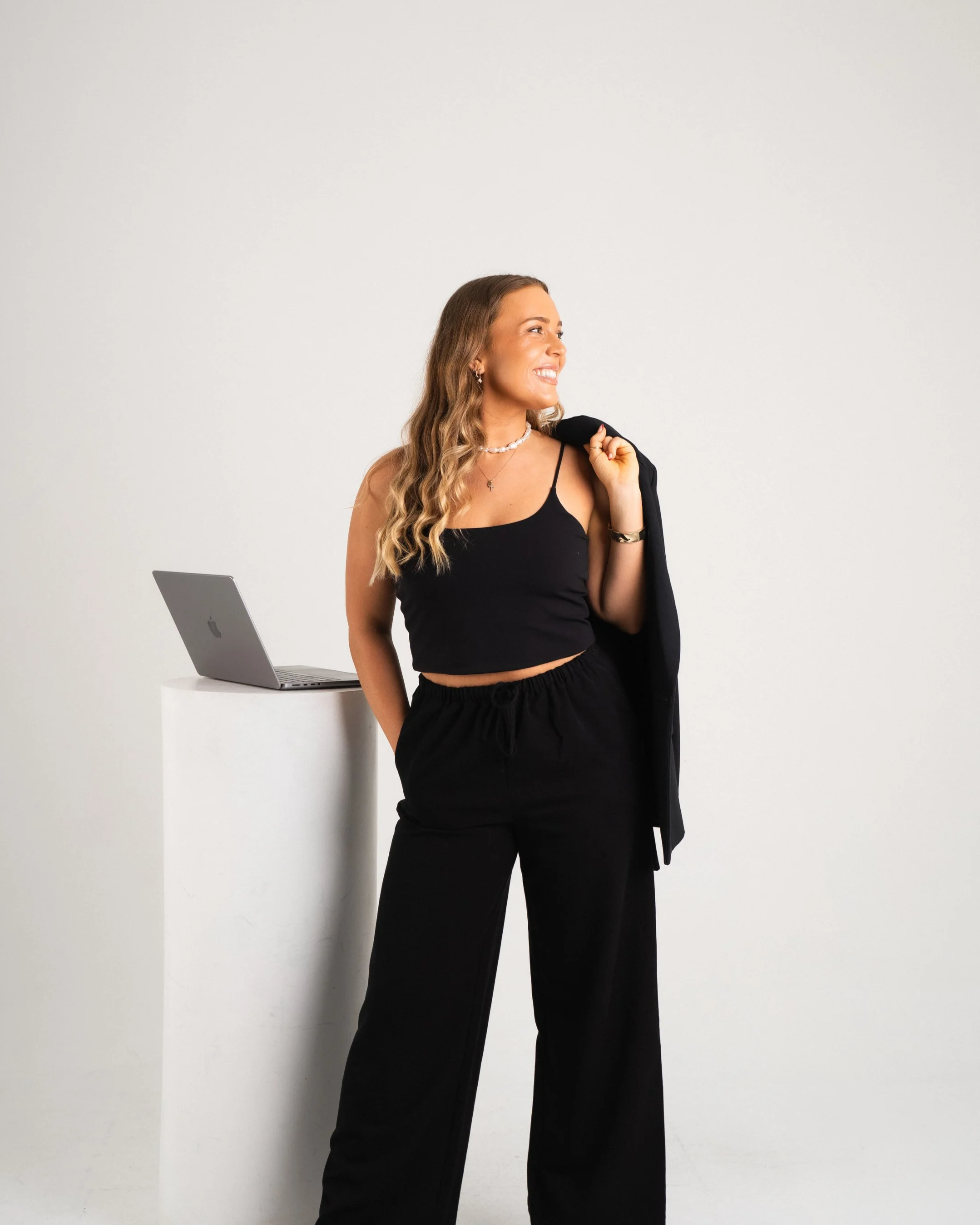 A woman with wavy blonde hair wearing a black crop top and black pants, holding a black blazer over her shoulder, standing next to a white pedestal with a silver laptop, smiling and looking to the side against a white background.