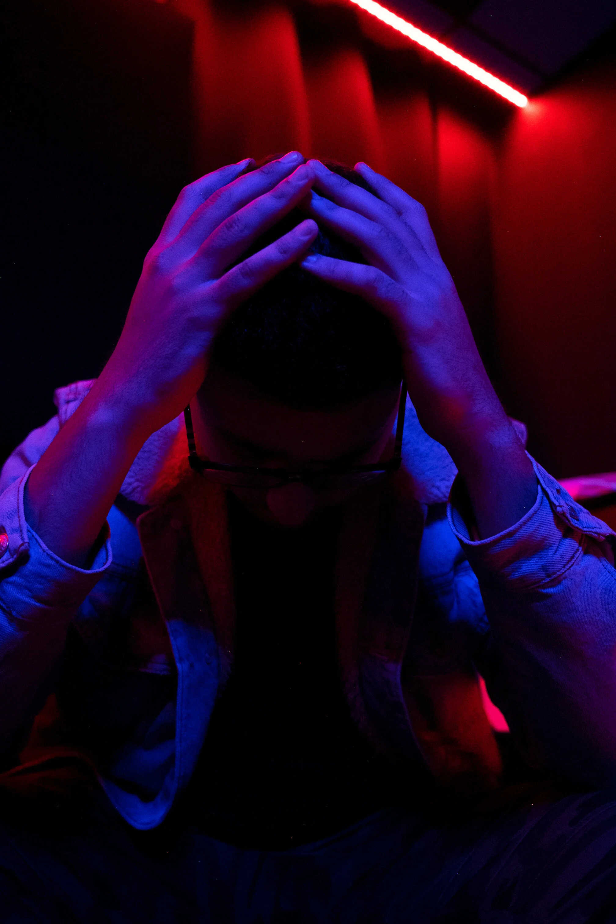Person in a dimly lit room with red and blue lighting, holding their head in their hands.