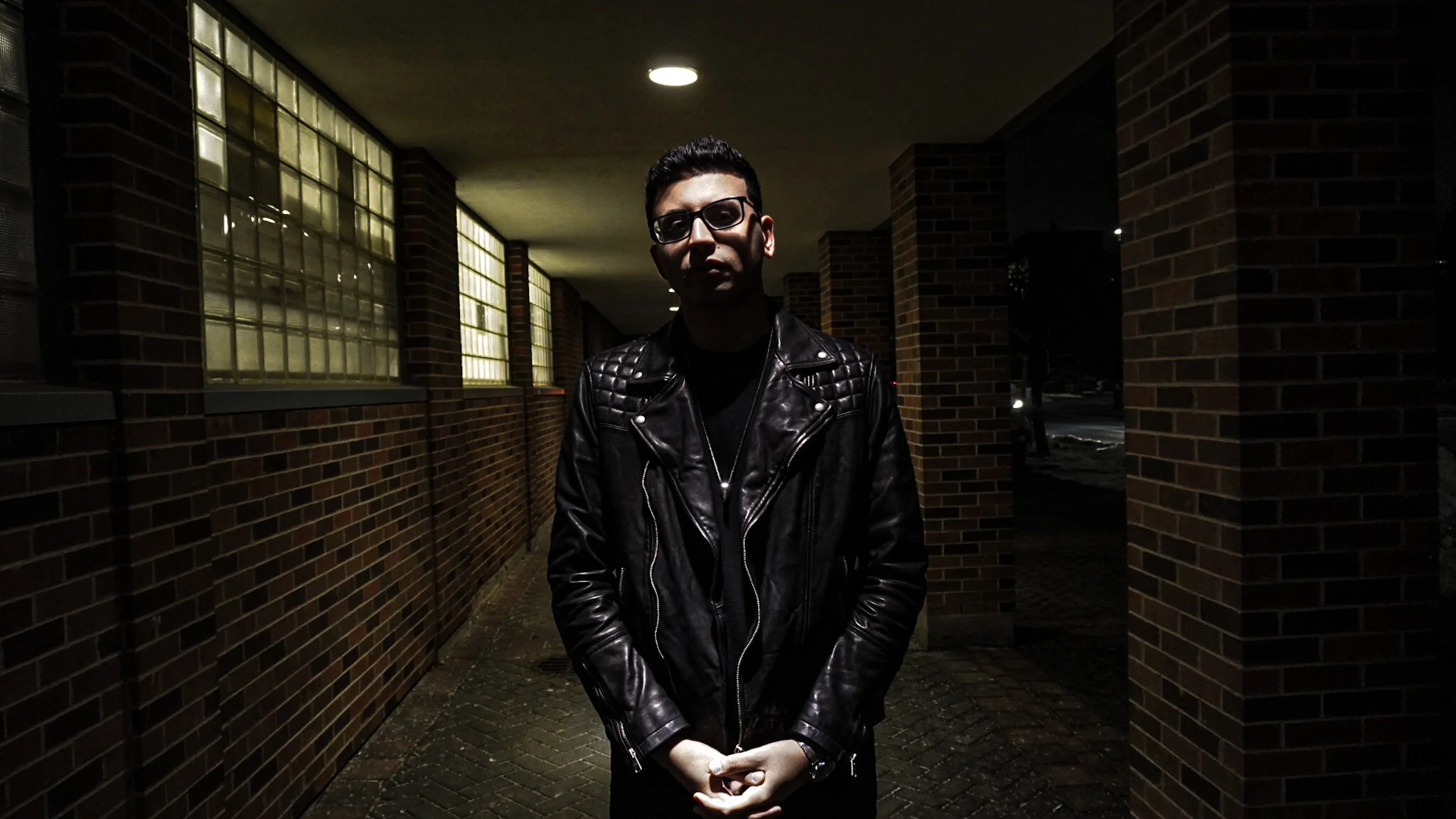 Person wearing a black leather jacket standing between brick walls at night.