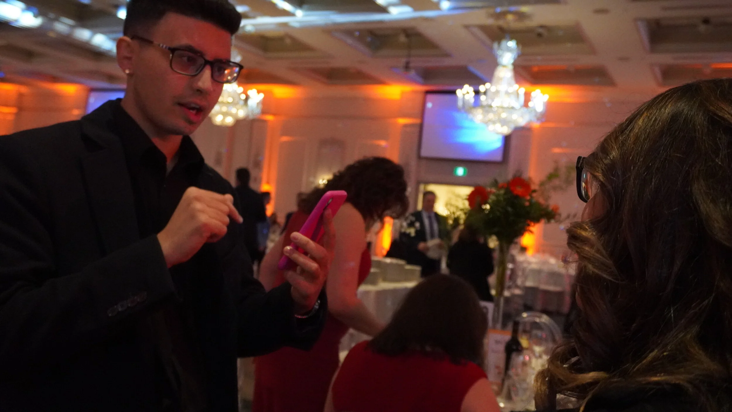 Man in a suit holding a smartphone and talking to a woman at an event in a decorated ballroom with chandeliers and orange lighting.