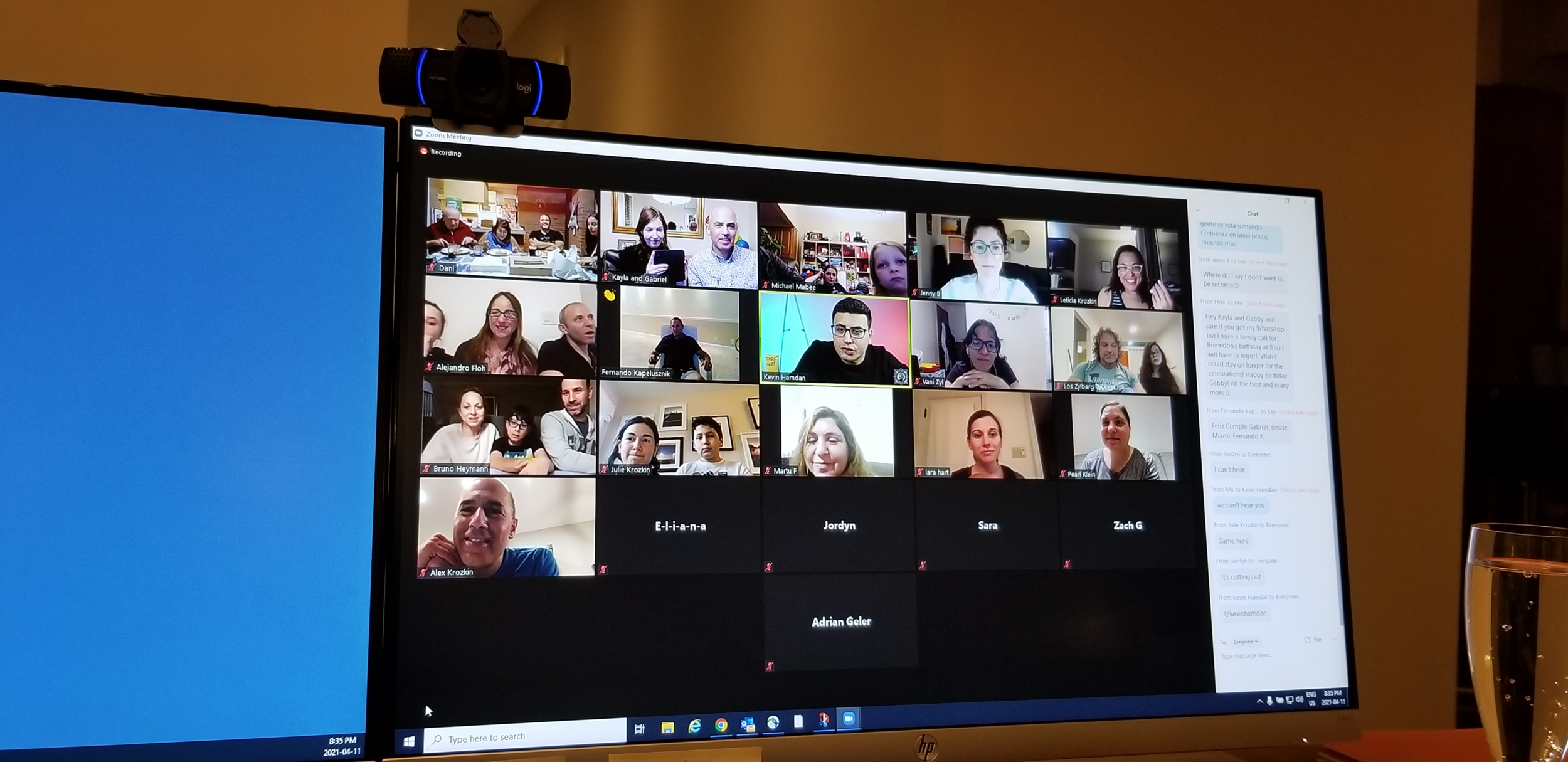 Computer screen displaying a Zoom meeting with multiple participants, showing their video feeds and names. A webcam is mounted on top of the monitor. Chat messages are visible on the side. A water glass is placed in front of the screen.