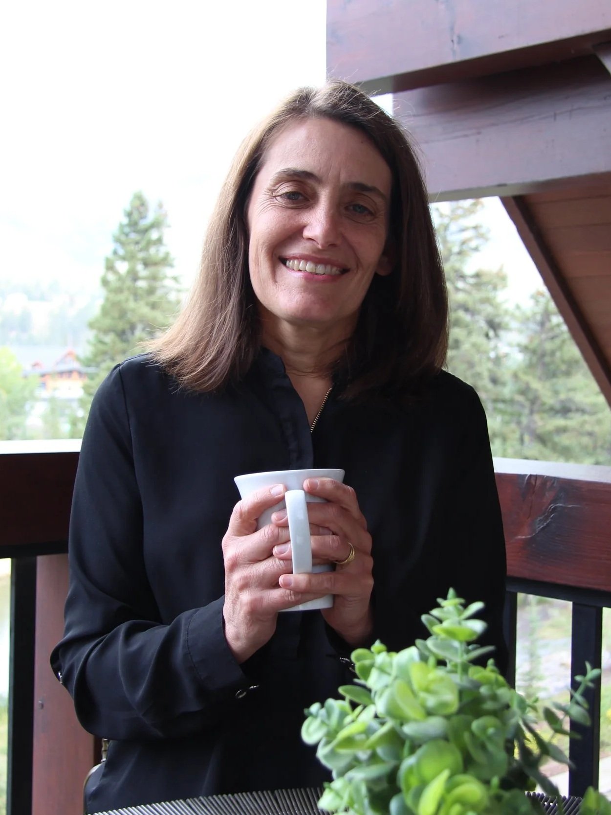 Cecile, a woman with shoulder-length brown hair smiling and holding a white coffee mug, sitting on a balcony with greenery and trees in the background.
