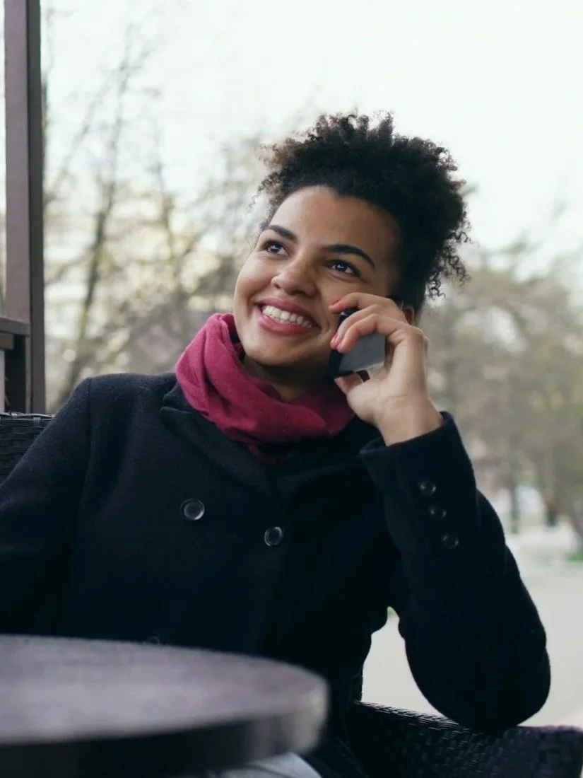 A woman with curly hair and a pink scarf smiling while talking on her phone at an outdoor cafe, with a drink on the table and trees outside.