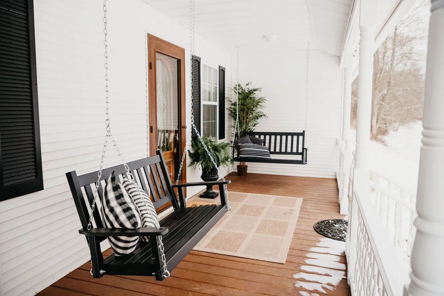 Porch with white siding, black window shutters, wooden door, black porch swing with striped pillows, potted plant, beige outdoor rug, snow outside.