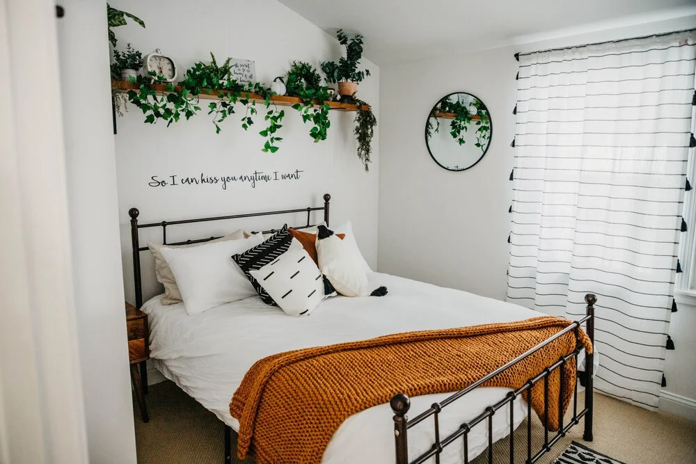 A bedroom with a black metal bed frame, white bedding, and decorative pillows. There is a mustard-colored knit blanket draped over the foot of the bed. A floating shelf with potted plants and decorative items is mounted on the wall above the bed, alo
