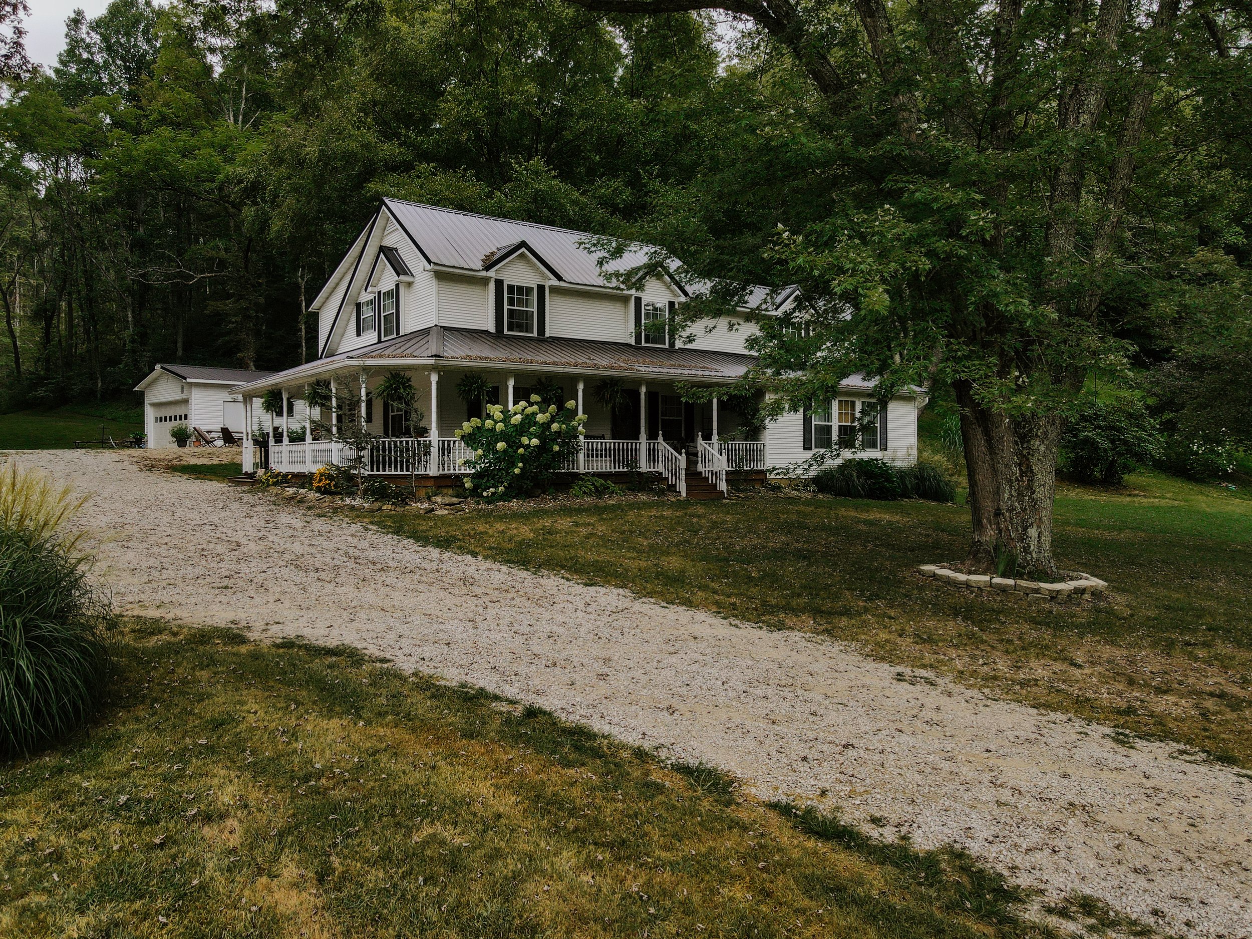 A large white house with a wraparound porch, surrounded by trees and greenery, with a gravel driveway in the front.