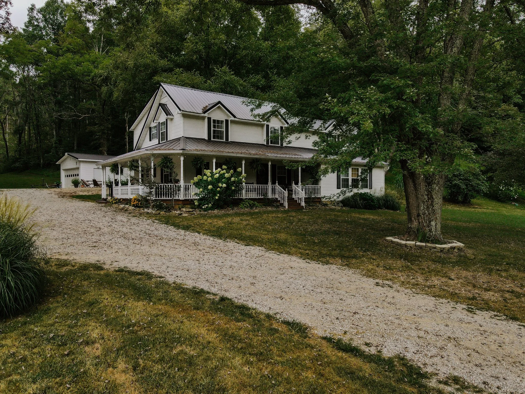A large white two-story house surrounded by trees, with a gravel driveway leading to the house. The house has a wraparound porch with white railings, and there are hanging plants and shrubs near the porch. There is a large tree with a stone border ar