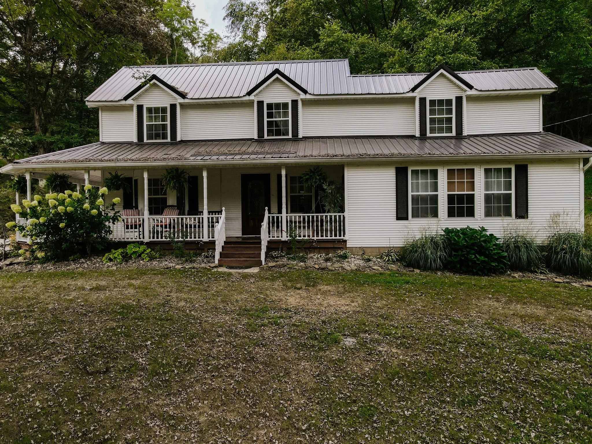 A two-story white house with black shutters and a metal roof, featuring a front porch with white railing, surrounded by trees and landscaping.