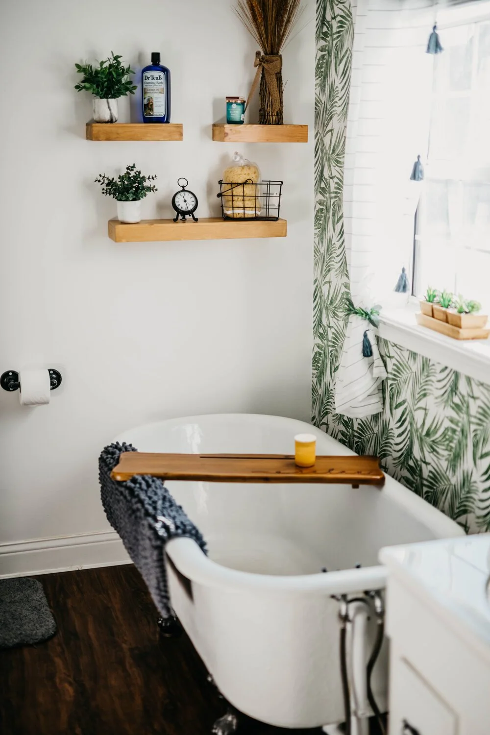 A bathroom with a white freestanding bathtub, a black and white towel draped over it, and a wooden bath tray holding a yellow candle. There are three wooden shelves on the wall with potted plants, a bottle of Dr. Teal's bath product, a small clock, a