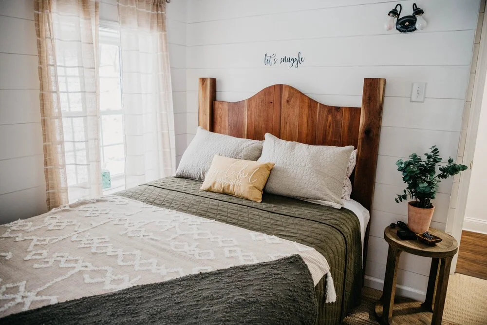 Cozy bedroom with white shiplap walls, a wooden headboard, and a bed with multiple pillows, including a decorative one that says 'joyful.' A small side table with a plant and remote controls is beside the bed.