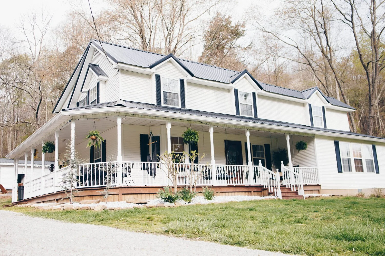 A two-story white house with black window shutters and a metal roof, surrounded by trees without leaves, with a front porch, potted plants, and a green lawn.