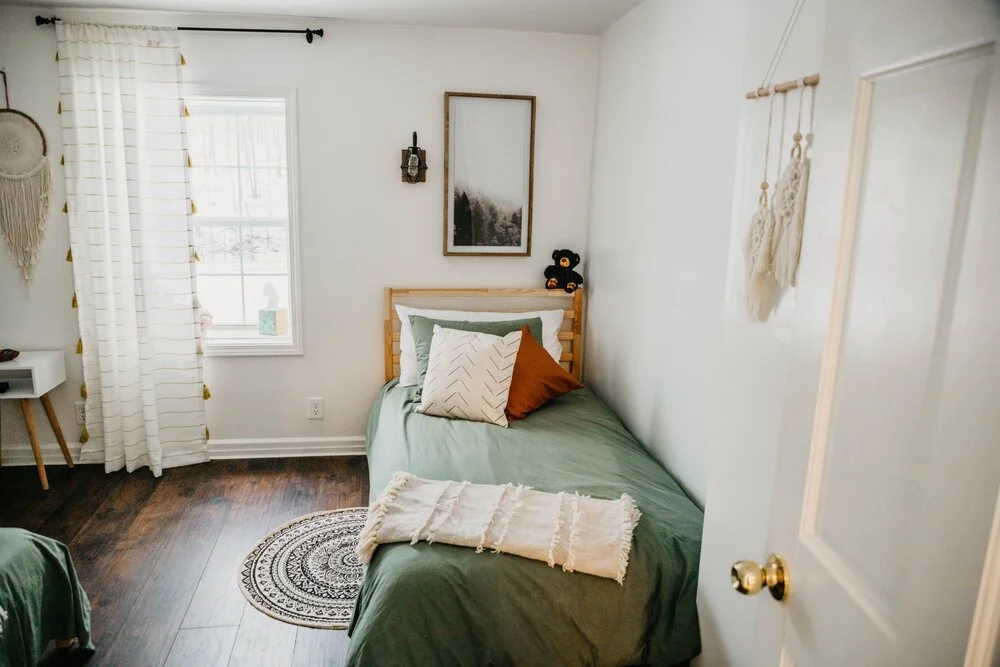 A cozy bedroom with a single bed, green bedding, and mixed pillows in beige, brown, and white. There are white curtains with tassels, a small white nightstand with wooden legs, a black-and-white framed photo, a small black wall hook, and wall hanging