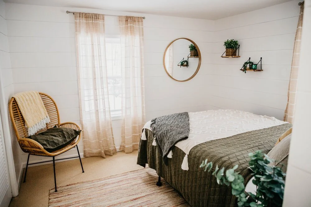 Cozy bedroom with white walls, a window with sheer beige curtains, a round mirror, a bed with white and gray bedding, a wicker chair with a blanket and pillow, and wall-mounted shelves with plants.