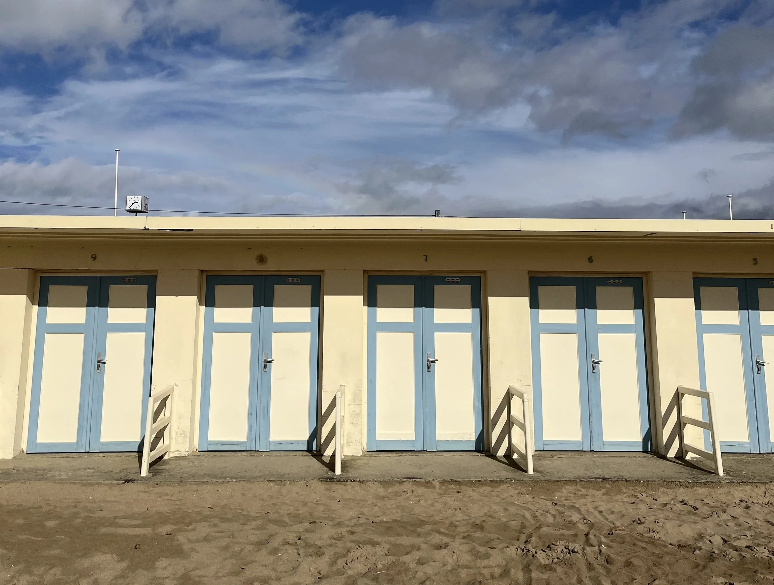 Empty beach huts with blue doors behind sandy ground under a cloudy sky.