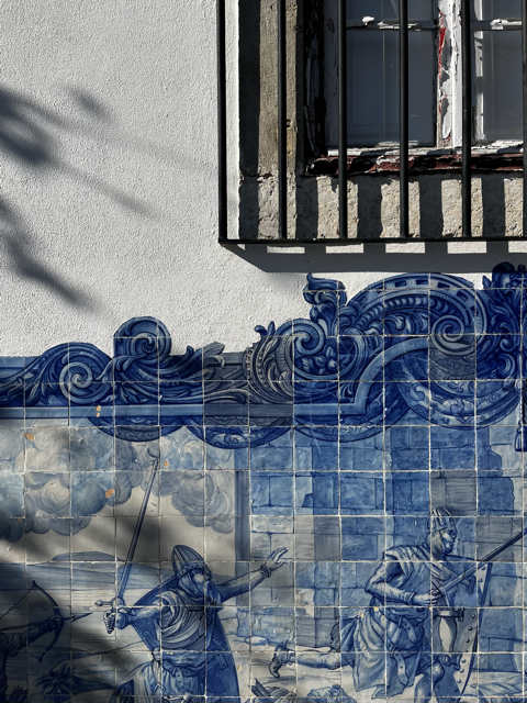 Close-up of a white exterior wall with a window barred with metal, and a decorative blue tile mural depicting a scene.