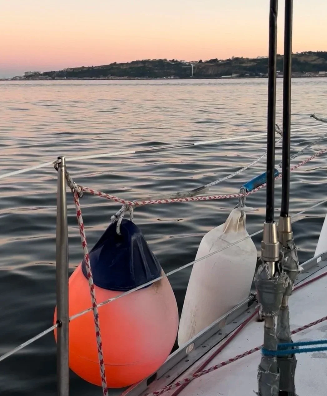 A boat's side with two floats, a blue one and a white one, attached to the side with ropes, with water and a distant landmass at sunset in the background.